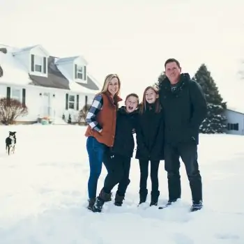 Family of four and farm dog stands in snow on farm with two story white house, white shop, and evergreen trees.
